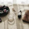 A serene, artistic, top-down shot of a diverse collection of sensory items arranged on a soft, textured cream-colored blanket. Items include a silk scarf, an ice cube melting in a glass bowl, a feather, a bottle of massage oil, and a journal with a pen. The lighting is warm and ambient, invoking a sense of calm, self-care, and intimate exploration without being explicitly sexual.