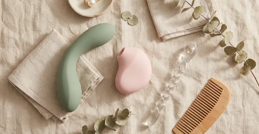 A tasteful, high-quality flat lay photography composition on a soft linen background. The image features an assortment of modern, abstract sexual wellness tools in pastel and neutral tones (sage green, soft pink, slate grey). Included are a curved silicone wand, a small air-pulse stimulator, and a sleek glass wand. The lighting is natural and warm, evoking a sense of wellness, safety, and modern sophistication suitable for a lifestyle blog in 2026.